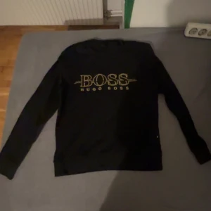 Svart långärmad tröja Hugo Boss - Svart långärmad tröja från Hugo Boss med stort gult BOSS-tryck på bröstet. Klassisk rund halsringning och mjukt material som passar perfekt till jeans eller joggers. Enkel och stilren design med fokus på loggan. Storlek XS