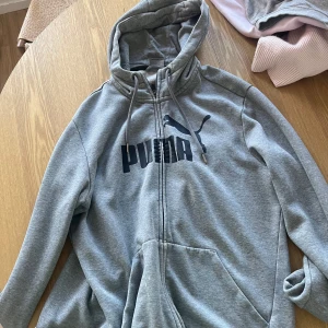 Grå hoodie från Puma med dragkedja - Säljer en grå hoodie från Puma med svart logga på bröstet. Tröjan har dragkedja, huva med snörning och stora fickor framtill. Koftan går lite över midjan, använd men har bra skick.