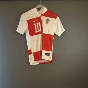 Kroatien Modric 10 Nike fotbollströja - Säljer en Kroatien fotbollströja från Nike med Modric och nummer 10 på ryggen. Tröjan har klassiskt rödvitt rutmönster, korta ärmar och broderat landslagsemblem på bröstet. Perfekt för dig som vill sticka ut på match eller i stan.