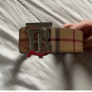 Burberry bälte med klassiskt rutmönster - Säljer ett Burberry bälte med det ikoniska beigea rutmönstret och en stor silvrig 'TB'-logga som spänne. Bältet är gjort i skinn och har detaljer i rött, svart och vitt. Perfekt accessoar för att lyfta din outfit med en lyxig touch.