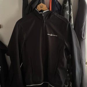 Svart vindjacka från Sail Racing - Svart vindjacka från Sail Racing med hel dragkedja framtill och vit logga på bröstet. Jackan har hög krage, två fickor och vita detaljer längs dragkedjan och nederkanten. Perfekt för blåsiga dagar och snygg till streetwear.