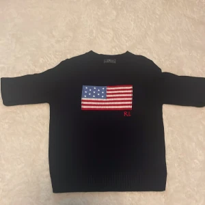 Svart stickad tröja Polo Ralph Lauren - Svart stickad tröja från Polo Ralph Lauren med amerikansk flagga broderad på bröstet och RL-detalj i rött. Klassisk rund halsringning och långa ärmar. Perfekt för dig som gillar stilrena plagg med en twist.