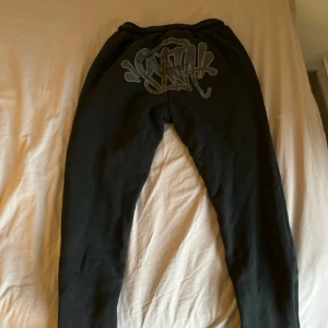 Syna World Tracksuit Black Edition - Central Cee - Svart hoodie från Syna World med grått graffiti-tryck på huvan och broderad text 'Forgive Me' på ärmen. Klassisk känguruficka framtill och mjukt material som känns skönt mot huden. Perfekt för dig som gillar streetwear och unika detaljer.