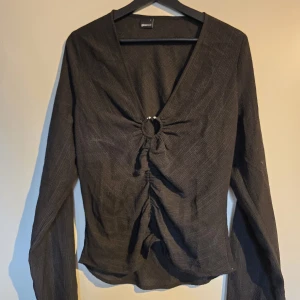 Svart långärmad topp med ringdetalj - Svart långärmad topp från Gina Tricot med v-ringning och snygg ringdetalj framtill. Toppen har rynkade detaljer längs magen och är tillverkad i ett lätt, något transparent tyg. Perfekt för dig som gillar trendiga och stilrena plagg. Är en storlek L men är som en M