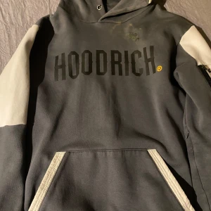 Grå och vit HOODRICH hoodie med dragkedja - Snygg hoodie från HOODRICH i grått med vita detaljer på ärmar och fickor. Stor logga framtill, huva och praktisk dragkedja på ena ärmen. Tillverkad i mjuk bomullsmix, perfekt för chill eller streetwear. Har en fäck som går å fixa om man är fiffig, kan inte hålla någon nål och fixa. Nypris 1000