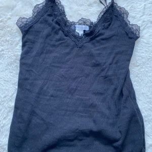 Svart spetslinnetopp från Amisu XS - Svart ribbad linnetopp från Amisu med smala axelband och v-ringad spetskant både fram och bak. Toppen är ärmlös och har en feminin look med fina spetsdetaljer längs urringningen. Perfekt att styla lager på lager eller som den är.