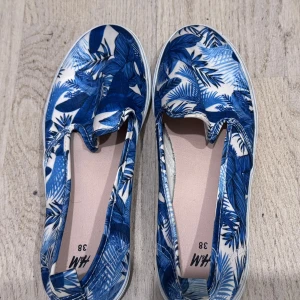 Blåvita slip-on sneakers H&M - Snygga slip-on sneakers från H&M i storlek 38 med blått och vitt bladmönster. Skorna har rund tå och platt sula, perfekta för en avslappnad stil. Tillverkade i textilmaterial med elastiska sidor. .