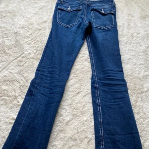 Blå bootcut jeans från Gina Tricot - Snygga blå jeans från Gina Tricot i modell bootcut med kontrastsömmar och fickor med lock och knapp bak. Klassisk femficksdesign och normal passform. Perfekta för dig som gillar en tidlös jeanslook med lite utsvängda ben.