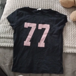 Svart t-shirt med rosa 77-tryck - HAR FLERA I DENNA STIL UPPLADDADE‼️‼️‼️‼️Svart t-shirt med stort rosa 77-tryck på bröstet. Klassisk rund halsringning och korta ärmar. Perfekt för en sportig och avslappnad stil. Gjord i mjukt bomullstyg som är skönt mot huden. Trycket är lite slitet, men det märks knappt!