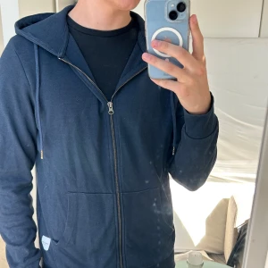 Jack & Jones Mörkblå hoodie  - Säljer en mörkblå hoodie från Jack & Jones Essentials i storlek M. Tröjan har dragkedja, huva med snörning och två stora fickor framtill. Perfekt basic med ribbade muddar och mjukt bomullsmaterial. Enkel och stilren design som funkar till allt.