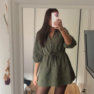 Olivgrön kort klänning  - Supersöt olivgrön kort klänning med v-ringning och knappar framtill. Klänningen har trekvartsärmar och ett skärp man kan knyta på olika sett. Passar perfekt året om! Använd fåtal gånger. I storlek M, men det går ju att spänna midjans som man själv vill. Från Zara.