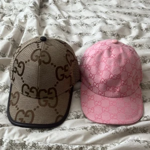 Gucci kepsar med GG-mönster - Två snygga Gucci kepsar med klassiskt GG-mönster. Ena kepsen är beige och brun med stora GG-loggor och svart kant, den andra är rosa med ljusrosa GG-mönster och rosa knapp. Båda har justerbar rem baktill och är tillverkade i bomullstyg med detaljer i läder.