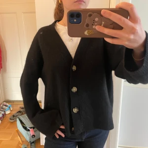 Svart kofta med stora knappar - Säljer en svart kofta med v-ringning och stora, snygga knappar framtill. Koftan har en loose passform och är stickad i ett mjukt material, perfekt för lager-på-lager. Långa ärmar och enkel, clean stil som funkar till allt.