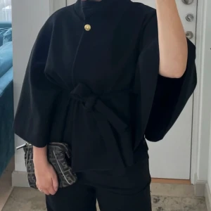 Svart ponchojacka med bälte och knapp - Snygg populär svart ponchojacka från Missäy- Blair med breda ärmar, hög krage och guldfärgad knapp framtill. Jackan har ett matchande tygbälte i midjan, så man kan anpassa hur man vill att den ska sitta. (Knappt använd, inga defekter).