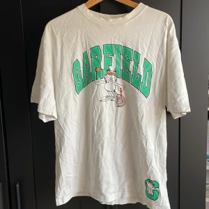 Vit Garfield t-shirt med tennistryck - Vit t-shirt med stort grönt Garfield-tryck på bröstet och en tecknad Garfield som spelar tennis. Extra detalj med ett grönt C och Garfield nere vid fållen. Skön passform och klassisk rund hals.