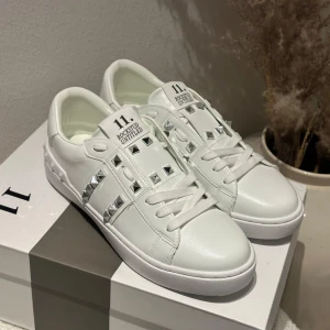 Valentino Rockstud vita sneakers - Snygga vita Valentino Rockstud sneakers i skinn med ikoniska silverfärgade nitar längs sidorna och hälen. Klassisk rund tå, platt sula och snörning framtill. Perfekta för dig som vill ha en clean look med edgy detaljer.