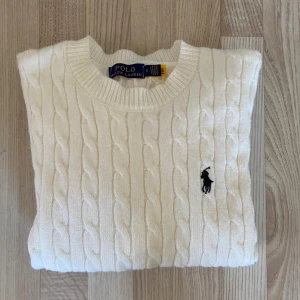 Vit kabelstickad tröja Polo Ralph Lauren - Vit kabelstickad tröja från Polo Ralph Lauren i 100% bomull. Klassisk rund halsringning, långärmad och med den ikoniska svarta loggan broderad på bröstet. Perfekt för en clean och stilren look.