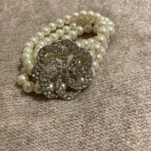 Pärlarmband med glittrig blomma - Snyggt armband med tre rader vita pärlor och en stor glittrig blomformad dekoration i silverfärg. Perfekt för dig som gillar att sticka ut med accessoarer som har extra bling och elegant vibe.
