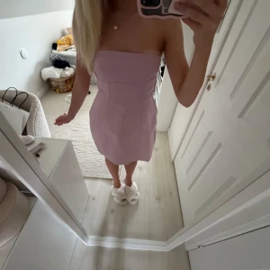 Ljuslila axelbandslös klänning med rosett - Superfin axelbandslös kort klänning i ljuslila/rosa med en stor rosett baktill. Klänningen har en figurnära passform och är sydd i ett slätt tyg som ger en stilren look. Perfekt för dig som vill sticka ut med en söt och trendig detalj.