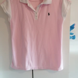 Rosa pikétröja från Polo Ralph Lauren XL - Snygg rosa pikétröja från Polo Ralph Lauren i 100% bomull. Klassisk krage och knäppning framtill, vita detaljer på krage och ärmar samt broderad logga på bröstet. Perfekt för en sportig och fräsch look.                              Tröjan är endast använd ett fåtal gånger, sedan har det hängt i garderoben. Vid bröstet har den två små fläckar. 