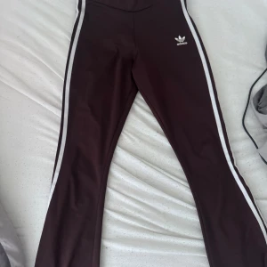 Bruna Adidas byxor med vita ränder XS - Snygga bruna Adidas byxor i stretchigt material med klassiska vita ränder längs sidorna och Adidas-logga på höften. Byxorna har hög midja och bootcut-snitt, perfekta för en sportig och trendig look.