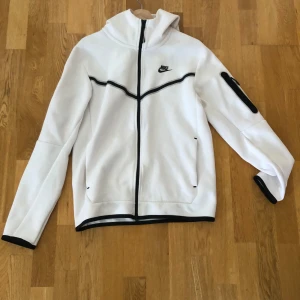 Vit Nike hoodie med svarta detaljer - Nike hoodie i vitt med svarta detaljer och dragkedja hela vägen. Tröjan har huva, lång ärm och en extra ficka med dragkedja på ena ärmen. Perfekt för dig som gillar sportig och clean stil.