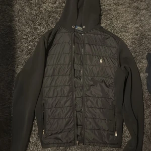 Svart quiltad jacka Polo Ralph Lauren - Svart quiltad jacka från Polo Ralph Lauren med huva och dragkedja. Jackan har broderad logga på bröstet, två fickor med dragkedja och snygga paneler i olika material. Perfekt för dig som vill ha en stilren och sportig look.
