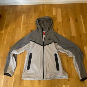 Grå vindjacka från Nike med huva - Storlek M, aldrig använt.Snygg grå vindjacka från Nike med tvåvägsdragkedja, huva och svarta detaljer. Jackan har två praktiska fickor med dragkedja och en sportig design med mörkare grå partier på axlar och ärmar. Perfekt för blåsiga dagar och streetwear-stil. 