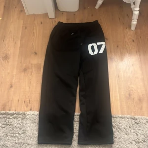 Svarta mjukisbyxor med tryck - Svarta mjukisbyxor med vitt tryck '07' på ena benet och snörning i midjan. Byxorna har en rak passform och är gjorda i mjukt material, perfekta för chill eller träning. Jag vet inte riktigt vart de är ifrån då hag fick de av en kompis och lappen var bortklippt. De har ibte kommit till användning och inte av min kompis eller så i väldigt bra skick.