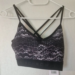 Svart mönstrad sport-bh från Röhnisch - Sport-bh från Röhnisch med svart bas och grått mönster, tunna korsade axelband och elastisk kant nedtill. Materialet är polyester och toppen har ficka för uttagbara kupor. Perfekt för träning med snygg design och flexibel passform.