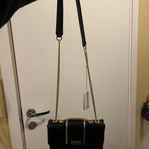 Svart axelväska från Guess med kedja - Snygg svart axelväska från Guess med guldfärgad kedja och logga framtill. Väskan har dekorativa sömmar och ett stilrent, rektangulärt format. Materialet är skinnimitation och axelremmen kombinerar kedja med läderdetalj för extra komfort och stil.