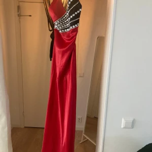 Röd långklänning med pärldetaljer - Exklusiv röd långklänning med svart och silverdetaljer. Stora pärlor och glittrande stenar handfästa av en sömmerska längs axel och slits ger en lyxig och elegant känsla. Asymmetrisk axel, elegant slits med svart mesh och glansigt material gör den perfekt för bal, bröllop, fest eller annan speciell afton.  Köpt i Esposa/ UAE för över 17 000 kr,  använd en gång, i nyskick. Size: 38-42  Litsn fläck på därav priset 