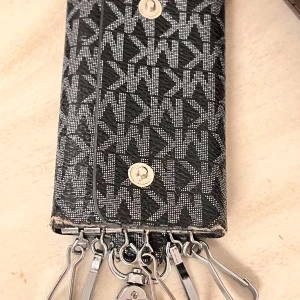Svart Michael Kors nyckelplånbok i skinn - Snygg svart nyckelplånbok från Michael Kors med silverfärgat MK-mönster. Plånboken har flera krokar för nycklar och stängs med tryckknappar. Tillverkad i tåligt skinn med metalliska detaljer och ett stilrent, grafiskt mönster.