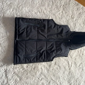 Svart dunväst med huva och dragkedja - Svart quiltad dunväst med stor huva och två fickor med dragkedja framtill. Västen har en rak passform och stängs med dragkedja hela vägen upp. Perfekt för lager-på-lager och chill stil.