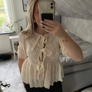 Krämvit blus med knyt från Gina Tricot - Supersöt krämvit blus från Gina Tricot med strukturmönster och tre knyt framtill. Blusen har volang nertill, puffiga halvlånga ärmar och en luftig, flowy passform. Perfekt för dig som gillar romantiska och trendiga detaljer.