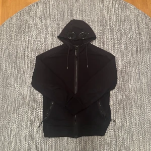  C.P. Company Hoodie Svart - Svart hoodie från C.P. Company med ikoniska goggles i huvan. Tröjan har dragkedja framtill, två fickor med dragkedja och justerbar huva med snörning. Tillverkad i mjuk bomull och har en clean, modern look som sticker ut.