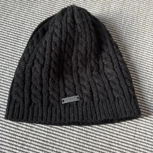 Lindberg - Svart kabelstickad mössa - Svart kabelstickad mössa från Lindberg med klassisk ribbad kant och snygg metallogo framtill. Tillverkad i mjukt och varmt material, perfekt för kyliga dagar. Passar huvudomfång 56-60 cm (strl 4). 