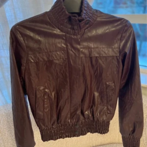 Vinröd skinnjacka från Zara Basic - Snygg vinröd skinnjacka från Zara Basic med cool bombermodell. Jackan har ribbade muddar vid krage, ärmslut och midja, samt dragkedja och tryckknappar framtill. Perfekt för dig som vill ha en stilren och trendig look.