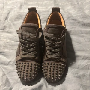 Gråa sneakers med nitar från Louboutin - Säljer ett par gråa sneakers från Louboutin med klassisk röd sula och coola silverfärgade nitar på tån men tappat två spikar men finns extra använda några gånger väldigt fint sick 