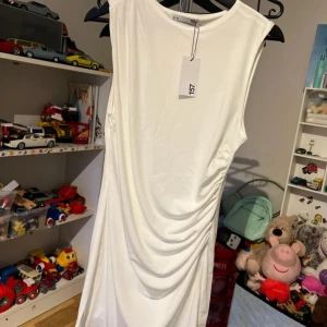 Vit ärmlös draperad klänning 157 - Snygg vit klänning från 157 med ärmlös design och draperad detalj på sidan. Klänningen har en rak passform och är tillverkad i ett mjukt, lätt material som ger en clean och modern look. Perfekt för dig som gillar minimalistisk stil.