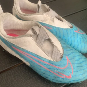 Nike Phantom GT2 fotbollsskor blå/rosa - Säljer ett par Nike Phantom GT2 fotbollsskor i blått och vitt med rosa detaljer och stor Nike-logga. Skorna har en modern design med snörning gömd under ett elastiskt band och mönstrad ovandel för extra grepp. Perfekta för dig som vill sticka ut på planen.