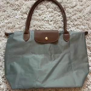 Grön handväska  - Snygg grön handväska med bruna handtag och lock i skinn. Väskan har klassisk design med guldfärgad knapp och rymligt huvudfack. Perfekt för dig som vill ha en stilren och praktisk väska till vardagen eller till skolan eller jobbet. 