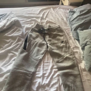 Grå mjukisbyxor från Nike med snörning - Grå mjukisbyxor från Nike med svart snörning i midjan och diskret Nike-logga på benet. Byxorna har en avslappnad passform och är tillverkade i mjukt bomullsmaterial. Perfekta för chill eller träning.