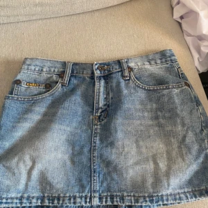 Blå jeanskjol från Cars Jeans - Snygg blå jeanskjol från Cars Jeans med klassisk femficksdesign, bälteshällor och gul logga på bakfickan. Kjolen har en rak passform och är i kort modell, perfekt till sneakers eller boots. Tidlös denim-look med slitna detaljer och kontrastsömmar.
