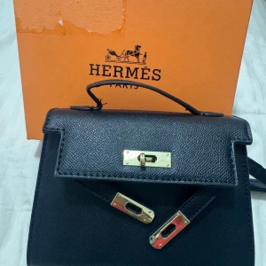 Svart handväska - Säljer en elegant svart handväska med guldfärgade detaljer. Väskan har en klassisk design med ett handtag och en stilren låsdetalj framtill. Perfekt för att ge en lyxig touch till din outfit. Kommer med boxen och ett extra band.