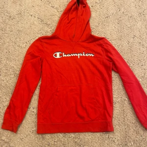 Röd hoodie från Champion - Säljer en röd hoodie från Champion med klassisk logga framtill. Tröjan har huva, lång ärm och en stor magficka. Perfekt för dig som gillar sportig och avslappnad stil.