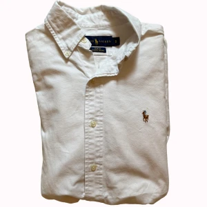 Vit skjorta från Ralph Lauren slim fit - Vit långärmad skjorta från Ralph Lauren i slim fit med klassisk krage och knappar framtill. Tillverkad i bomull med broderad polospelare på bröstet. Stretch Oxford-tyg ger extra komfort och snygg passform. Perfekt för dig som gillar stilrena och tidlösa plagg.