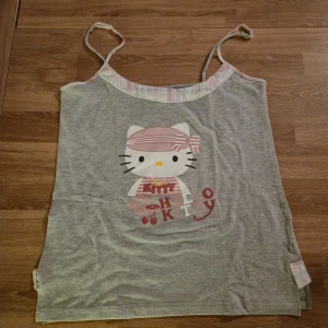 Grå Hello Kitty linnetopp L - Supersöt grå linnetopp med Hello Kitty-tryck framtill och tunna axelband. Toppen har en ljusrosa och vitrandig kant upptill och knappdetalj i sidan. Tillverkad i mjuk bomull med lite stretch för extra komfort. Varan är helt oanvänd med etiketten kvar.