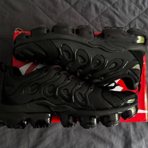 Nike Air Vapormax Plus Svarta Sneakers - Nike Air Vapormax Plus i helsvart med futuristisk design och synlig luftdämpning i sulan. Ovandelen är i syntet och textil med vågformade detaljer och snörning. Perfekta för dig som vill ha en modern och sportig look.