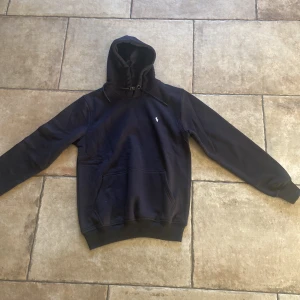 Svart hoodie från Polo Ralph Lauren - Svart hoodie från Polo Ralph Lauren med klassisk huva och snörning. Tröjan har en broderad vit logga på bröstet och en stor magficka. Tillverkad i mjuk bomull, perfekt för chill dagar. Snygg och enkel att matcha med det mesta.